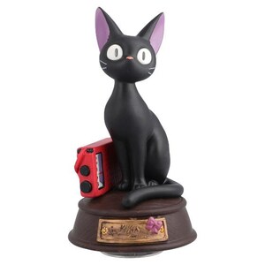 May include: A black cat figurine with purple-lined ears, white eyes, and whiskers sits atop a brown base. A red box is next to the cat. The base has a decorative plaque with a flower.