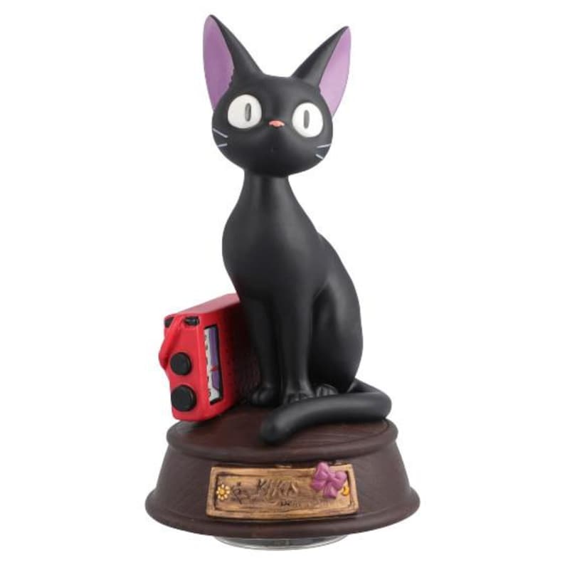 May include: A black cat figurine with purple-lined ears, white eyes, and whiskers sits atop a brown base. A red box is next to the cat. The base has a decorative plaque with a flower.