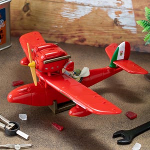 May include: A bright red seaplane model featuring a yellow propeller, a pilot figure in the cockpit, and a tail fin decorated with the Italian flag colors of green, white, and red. The aircraft is positioned on a stand with tools nearby.