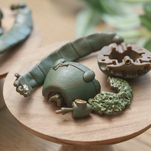Peut inclure: Divers &eacute;l&eacute;ments de jouets vert olive, dont une t&ecirc;te, un bras et d'autres composants, sont dispos&eacute;s sur une surface en bois. Le bras pr&eacute;sente un petit d&eacute;tail floral. Les pi&egrave;ces semblent provenir d'un jouet plus grand.