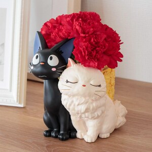 Puede incluir: Una maceta decorativa con dos gatos esculpidos, uno negro con ojos grandes y orejas bordeadas de azul, y otro blanco con ojos cerrados y pelaje esponjoso. Un ramo de claveles rojos est&aacute; dispuesto en el recipiente amarillo detr&aacute;s.