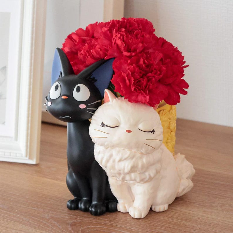 Puede incluir: Una maceta decorativa con dos gatos esculpidos, uno negro con ojos grandes y orejas bordeadas de azul, y otro blanco con ojos cerrados y pelaje esponjoso. Un ramo de claveles rojos est&aacute; dispuesto en el recipiente amarillo detr&aacute;s.