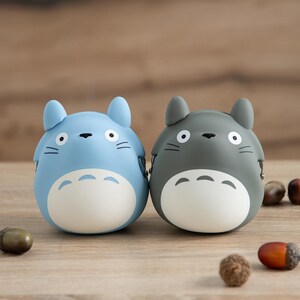 May include: Two cartoon-style figurines, one light blue and one gray, with white bellies and black facial details. The figurines are round with pointed ears, resembling a popular animated character. Acorns are scattered around the figurines on a wooden surface.