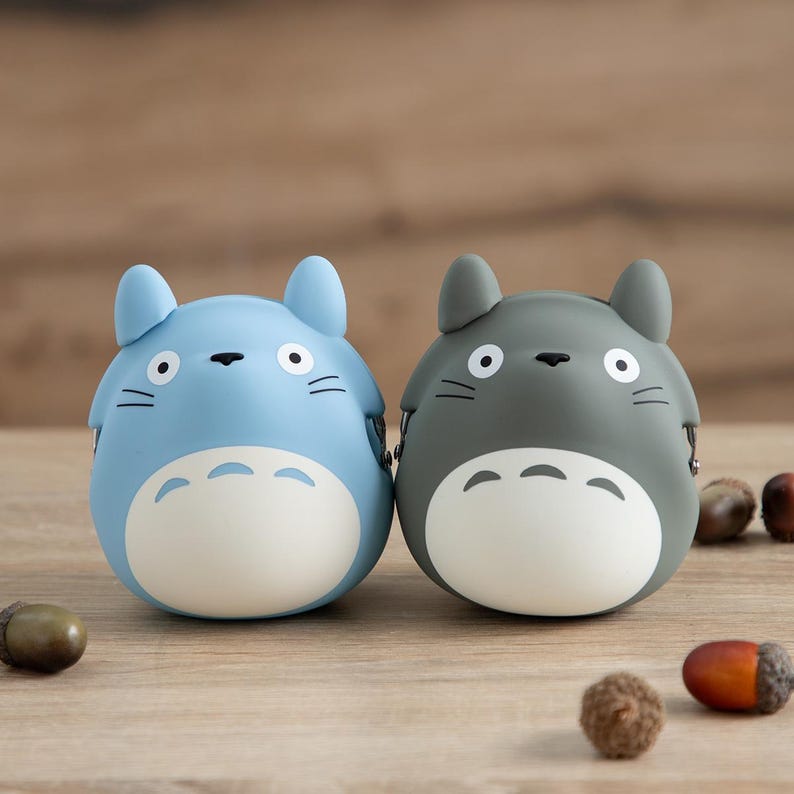 May include: Two cartoon-style figurines, one light blue and one gray, with white bellies and black facial details. The figurines are round with pointed ears, resembling a popular animated character. Acorns are scattered around the figurines on a wooden surface.