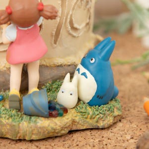 Puede incluir: Una figura que presenta un personaje Totoro azul con un personaje blanco m&aacute;s peque&ntilde;o y una ni&ntilde;a con un vestido rosa. La escena incluye un cubo azul volcado con objetos coloridos, sobre una base de hierba. Probablemente un art&iacute;culo de colecci&oacute;n o decorativo.