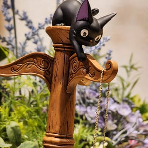 May include: A black cat figurine with purple inner ears perches on a decorative wooden stand. The stand is brown with ornate carvings and holds a delicate gold-colored necklace. The cat has large, wide eyes.