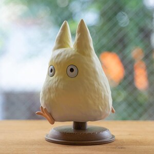 May include: A light yellow figurine of a character with large eyes and pointed ears, resembling a creature from an animated film. The figurine is mounted on a brown base with a wooden texture, and has small arms and feet.