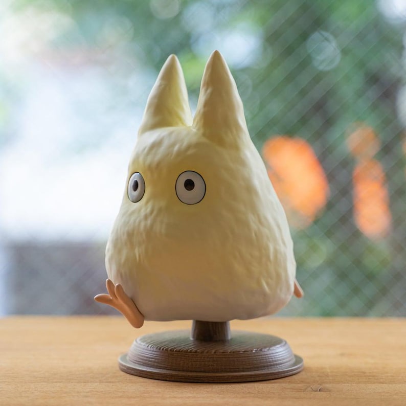 May include: A light yellow figurine of a character with large eyes and pointed ears, resembling a creature from an animated film. The figurine is mounted on a brown base with a wooden texture, and has small arms and feet.