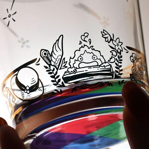 May include: A clear glass with a gold rim and black illustrations. The illustrations include a character, flowers, and other decorative elements. The glass has a colorful, striped design at the bottom, with red, blue, green, and purple hues.