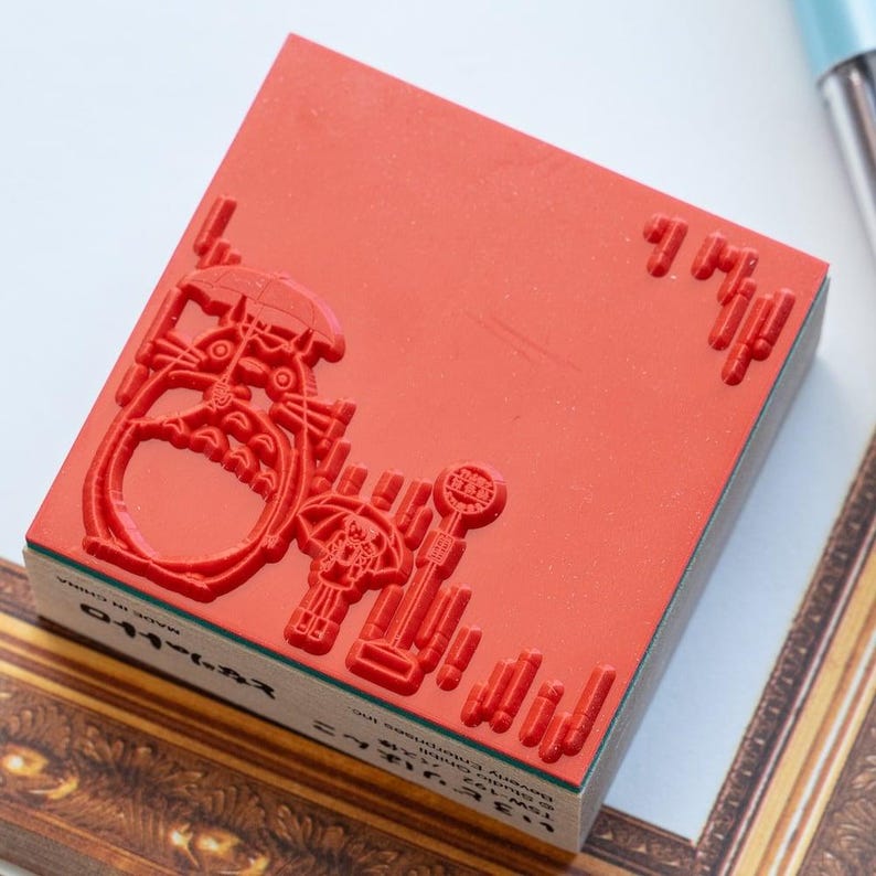 Op de afbeelding: Een vierkante rode rubberen stempel op een houten blok. De stempel heeft een verhoogd ontwerp van een groot wezen met een paraplu, een kleiner figuur met een paraplu, een bushaltebord en regenstrepen. Tekst op de zijkant leest Studio Ghibli.