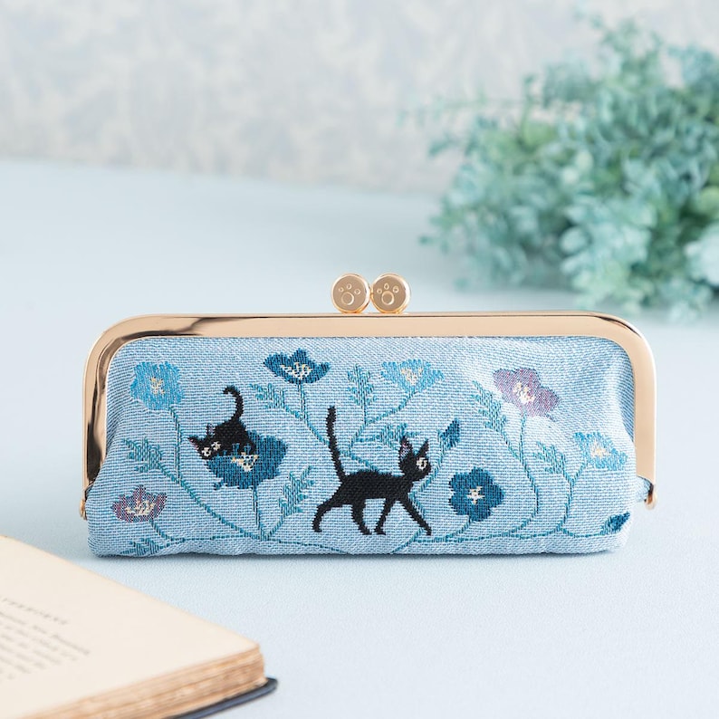 May include: A light blue fabric clutch with a gold metal frame and two gold metal paw print accents. The clutch features an embroidered design of black cats walking through a field of blue flowers.