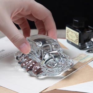 May include: A clear glass car-shaped paperweight with intricate detailing. The paperweight is held by a hand, and sits on a piece of paper. An ink bottle is in the background.