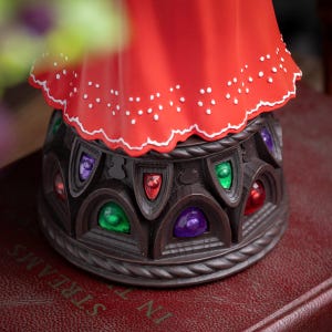 Puede incluir: Una figura decorativa con una parte superior roja y una base marr&oacute;n. La parte superior roja tiene un borde festoneado blanco con puntos blancos. La base presenta coloridos acentos de joya en verde, morado y rojo. La figura est&aacute; sobre un libro rojo.