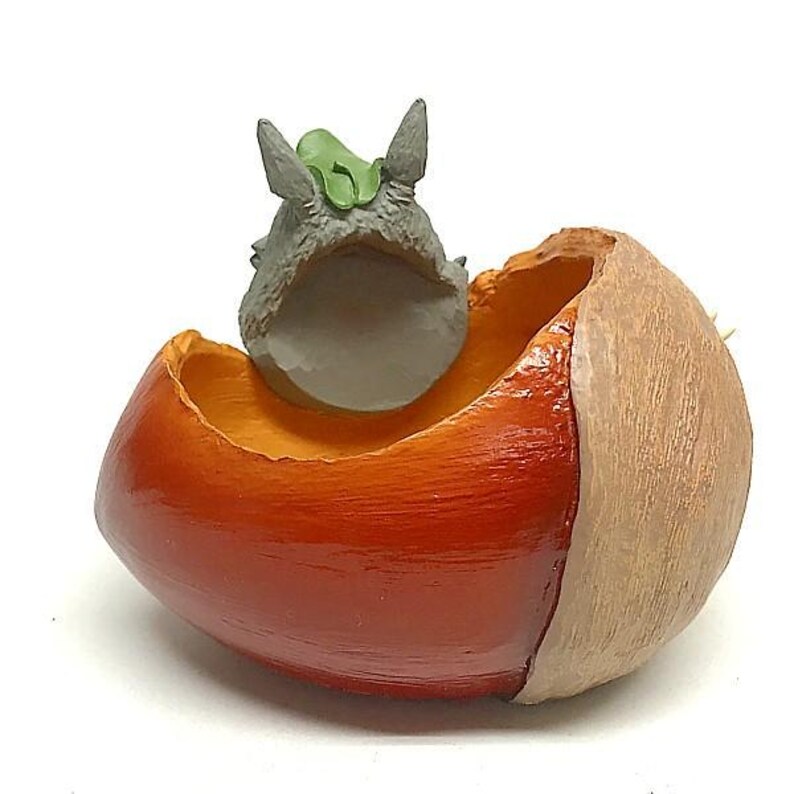 K&ouml;nnte beinhalten: Eine skurrile Figur, die ein graues Wesen mit spitzen Ohren und einem gr&uuml;nen Blatt zeigt, das in einem eichelf&ouml;rmigen Beh&auml;lter sitzt. Der Beh&auml;lter ist orange und braun, mit gl&auml;nzendem Finish. Die Eichel ist ca. 5 cm lang.