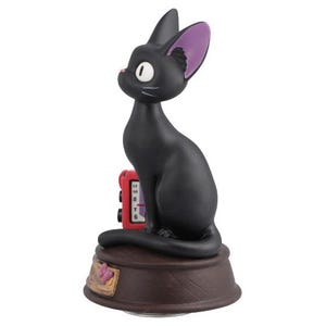 May include: A black cat figurine with purple inner ears and white eyes sits atop a brown base. A red clock is behind the cat. The figurine has a cartoonish design.