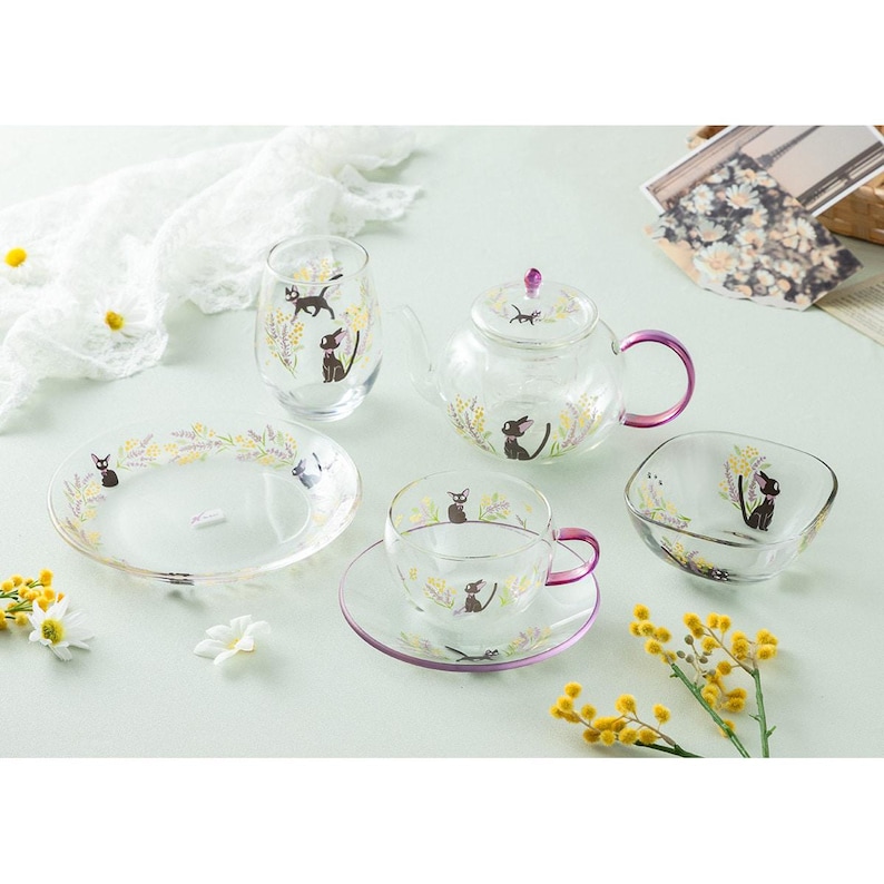 Op de afbeelding: Een theeservies van helder glas bestaande uit een theepot, een theekop met schotel, een klein kommetje, een glas en een bord. Elk item is versierd met illustraties van een zwarte kat en gele bloemtakjes. De theepot en theekop hebben paarse handvatten.