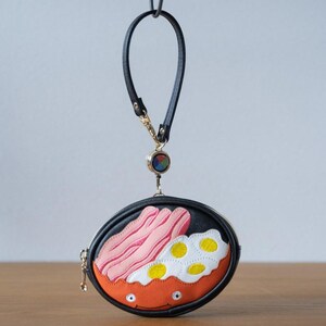 May include: Black, round coin purse with a breakfast-themed design. The purse has a zip closure, a black wrist strap, and a gold-coloured clasp. The design includes bacon, fried eggs, and an orange bowl.