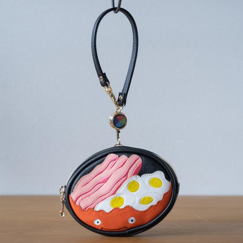 May include: Black, round coin purse with a breakfast-themed design. The purse has a zip closure, a black wrist strap, and a gold-coloured clasp. The design includes bacon, fried eggs, and an orange bowl.