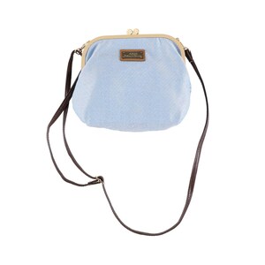 May include: A light blue, denim-style crossbody bag with a kiss-lock closure. The bag has a brown leather strap and a small rectangular tag with text. The frame is gold-colored.