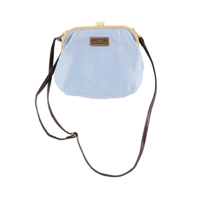 May include: A light blue, denim-style crossbody bag with a kiss-lock closure. The bag has a brown leather strap and a small rectangular tag with text. The frame is gold-colored.
