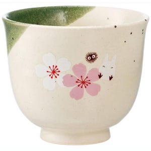 May include: Cream-colored ceramic teacup with a green rim. The cup features white and pink cherry blossom designs and a cartoon character illustration. The cup has a wide, rounded shape and a small footed base.