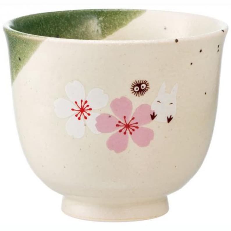 May include: Cream-colored ceramic teacup with a green rim. The cup features white and pink cherry blossom designs and a cartoon character illustration. The cup has a wide, rounded shape and a small footed base.