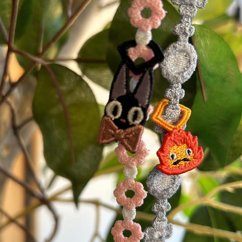 Op de afbeelding: Geborduurde bedelarmband met een zwarte kat, een bruine vlinderdas en een vurige figuur. De armband heeft een zilveren ketting en roze bloemaccenten. Achtergrond met groene bladeren.
