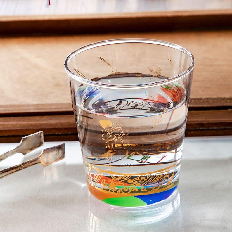 May include: A clear glass with a colorful, ornate design at the base and a gold illustration of a musician. The glass is filled with a clear liquid, possibly water. A pair of silver tongs rests on the side.