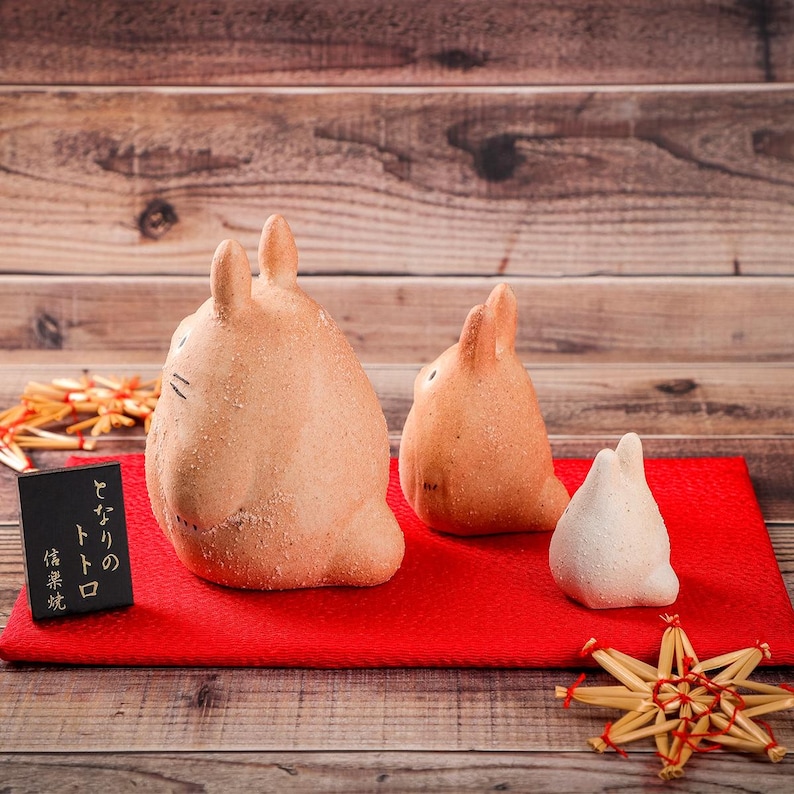 May include: Three light brown, bunny-shaped figurines with black facial details sit on a red surface. A small black sign with Japanese text is on the left. The background is a wooden surface.