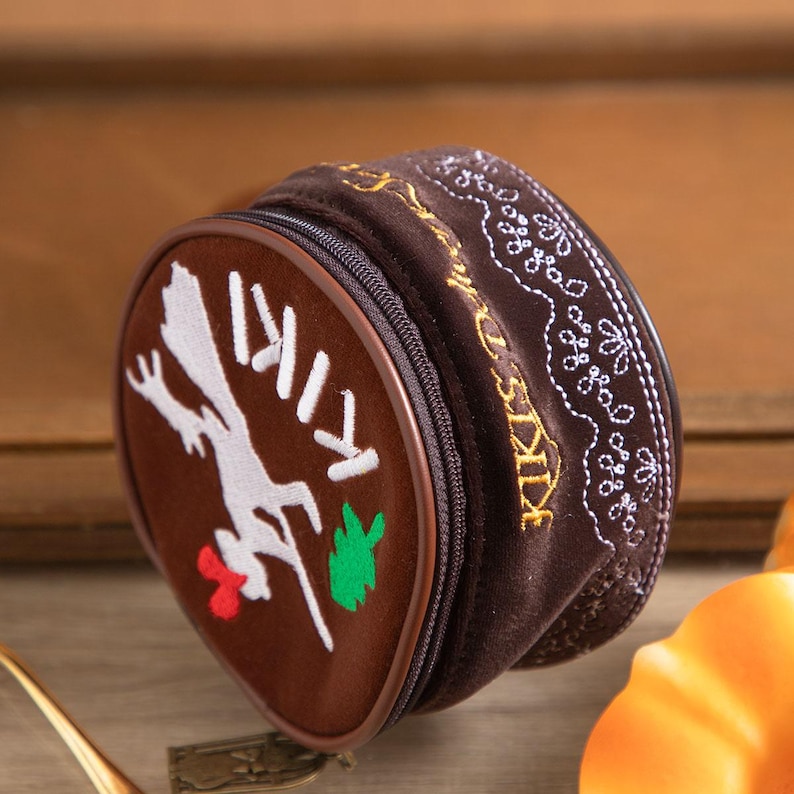May include: A brown, zippered pouch with the word "KIKI" and a white rooster graphic. The pouch has a brown border and gold and white embroidered details. The rooster has a red comb and a green leaf.