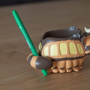 May include: A brown and orange pen holder shaped like a cartoon character, with a green pen inserted. The pen holder has a detailed design with a rounded shape and a textured surface. The pen is green with white polka dots.