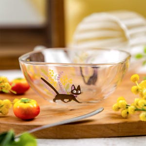 May include: A clear glass bowl featuring a graphic illustration of a black cat with a pink collar, surrounded by yellow mimosa flowers and purple sprigs. The bowl has a smooth, rounded rim and a transparent body that shows the design clearly.