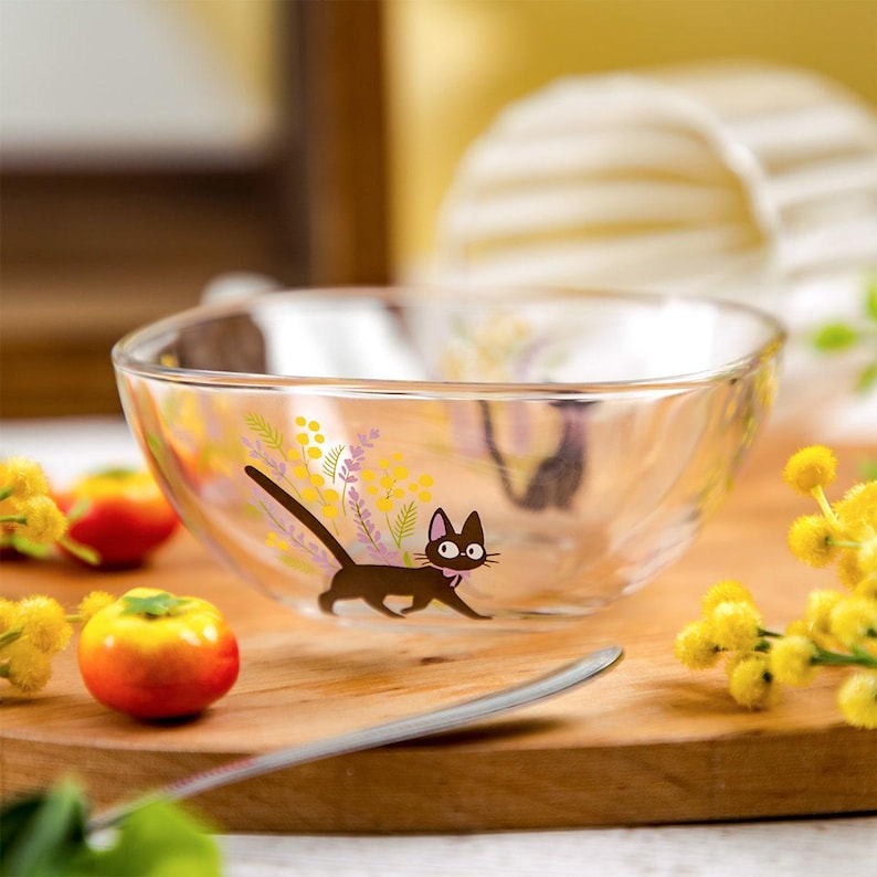 May include: A clear glass bowl featuring a graphic illustration of a black cat with a pink collar, surrounded by yellow mimosa flowers and purple sprigs. The bowl has a smooth, rounded rim and a transparent body that shows the design clearly.