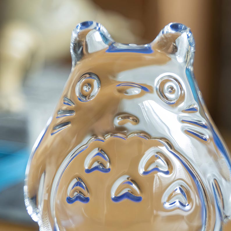 May include: A clear glass figurine in the shape of a cartoon character with large eyes, pointed ears, and a round body. The figurine has a reflective surface, and the details are etched into the glass. The character's belly features several heart-shaped designs.