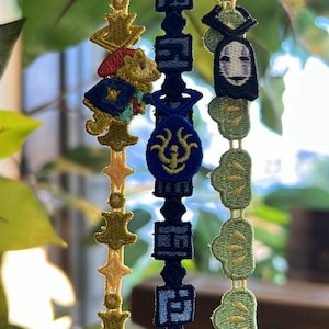 K&ouml;nnte beinhalten: Nahaufnahme von drei bestickten B&auml;ndern mit verschiedenen Designs. Ein Band zeigt goldene Sterne und Kronen, ein anderes blaue geometrische Formen und einen Anh&auml;nger, und das dritte einen No-Face-Charakter und gr&uuml;ne Blattformen.
