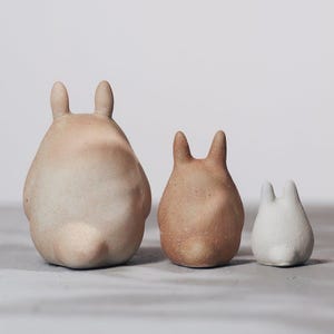 May include: Three ceramic bunny figurines in varying sizes and colors. The largest is a light beige, the middle one is a warm brown, and the smallest is white. Each has upright ears and a rounded body, perfect for home decor.