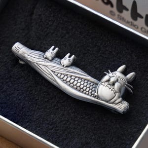 Może przedstawiać: Srebrna metalowa broszka z motywem Totoro i dwoma mniejszymi Totoro na kłodzie. Broszka jest prezentowana w czarnym pudełku, z logo Studio Ghibli widocznym na krawędzi. Broszka ma szczegółowe tekstury.