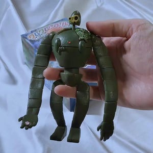 Peut inclure: Figurine de robot articul&eacute; vert olive avec une petite t&ecirc;te et des membres segment&eacute;s. La figurine a un petit d&eacute;tail en forme de c&oelig;ur sur sa poitrine et un petit &eacute;l&eacute;ment de t&ecirc;te dor&eacute;. La figurine est tenue dans une main.