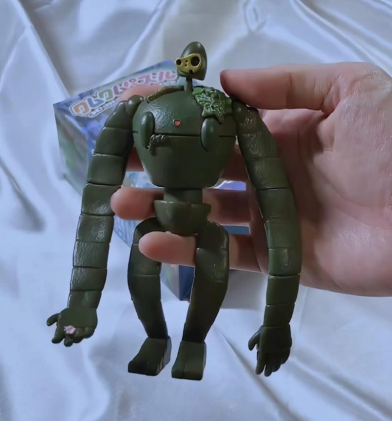 Peut inclure: Figurine de robot articul&eacute; vert olive avec une petite t&ecirc;te et des membres segment&eacute;s. La figurine a un petit d&eacute;tail en forme de c&oelig;ur sur sa poitrine et un petit &eacute;l&eacute;ment de t&ecirc;te dor&eacute;. La figurine est tenue dans une main.