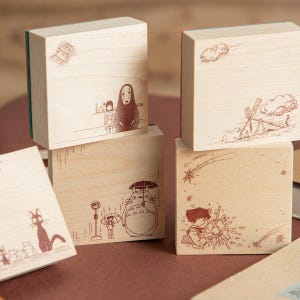 May include: A collection of four square wooden blocks featuring brown line art illustrations of various animated characters and scenes, including a large creature with an umbrella, a hooded figure, and a cat, all with a natural light wood finish.