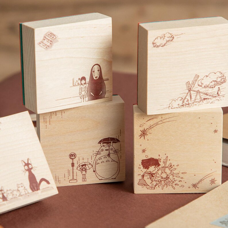 May include: A collection of four square wooden blocks featuring brown line art illustrations of various animated characters and scenes, including a large creature with an umbrella, a hooded figure, and a cat, all with a natural light wood finish.