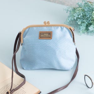 May include: A light blue denim-style crossbody bag with a kiss-lock closure and a brown strap. The bag features a gold-colored frame and a small rectangular label with text. The bag is a stylish accessory.