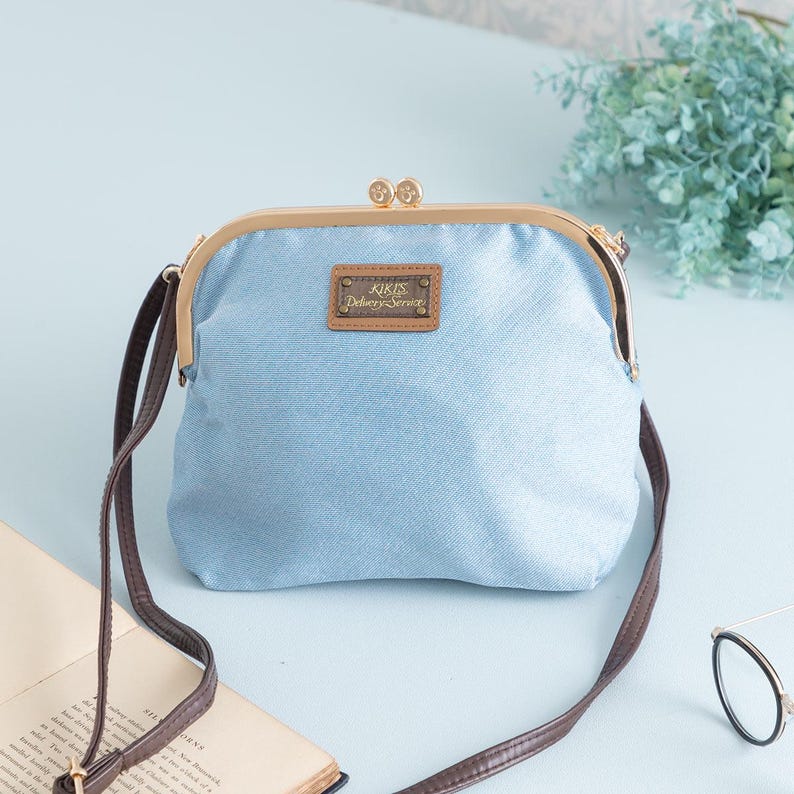 May include: A light blue denim-style crossbody bag with a kiss-lock closure and a brown strap. The bag features a gold-colored frame and a small rectangular label with text. The bag is a stylish accessory.