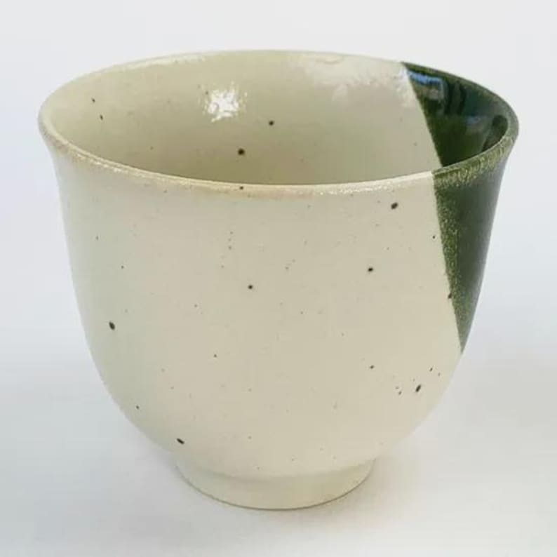 May include: A ceramic teacup with a speckled off-white exterior and a diagonal section of dark green glaze. The cup has a rounded shape and a small, circular base. The interior is off-white.