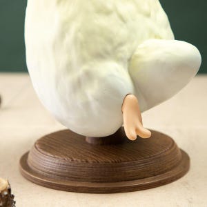 May include: A light yellow and white sculpture of a bird's rear end, with a small, tan foot extending from the bottom. The sculpture is mounted on a round, wooden base with a dark brown finish. The bird's form has a smooth, textured appearance.
