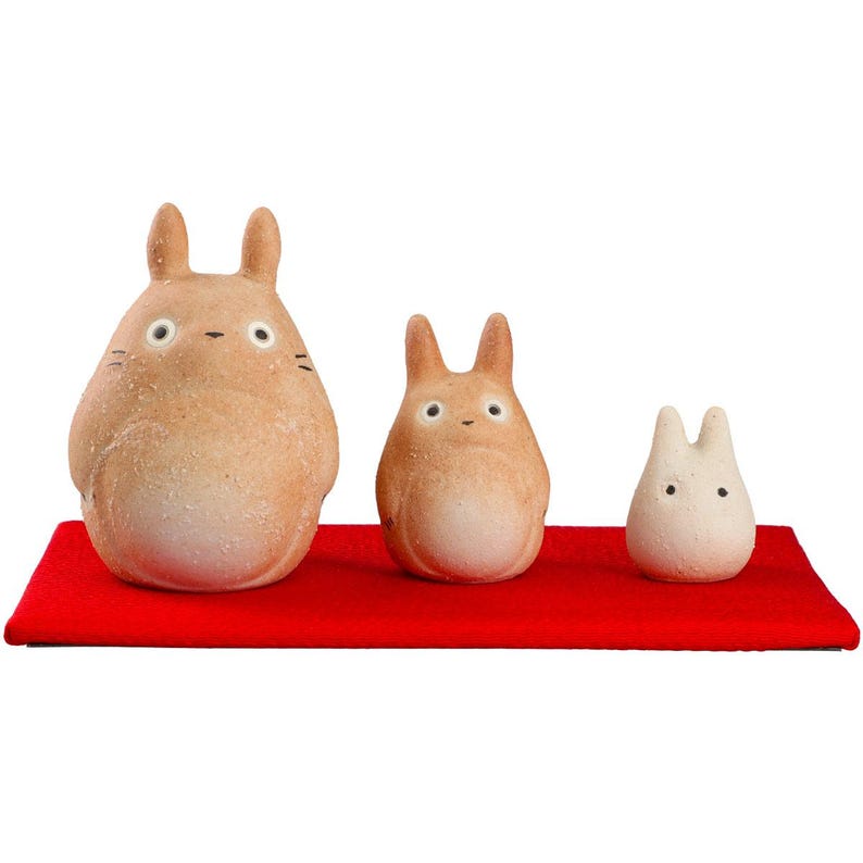 May include: Three tan-colored Totoro figurines of varying sizes are displayed on a red rectangular base. The largest figure has a round body and pointed ears, with small black eyes and whiskers. The other two figures are smaller, with similar features.