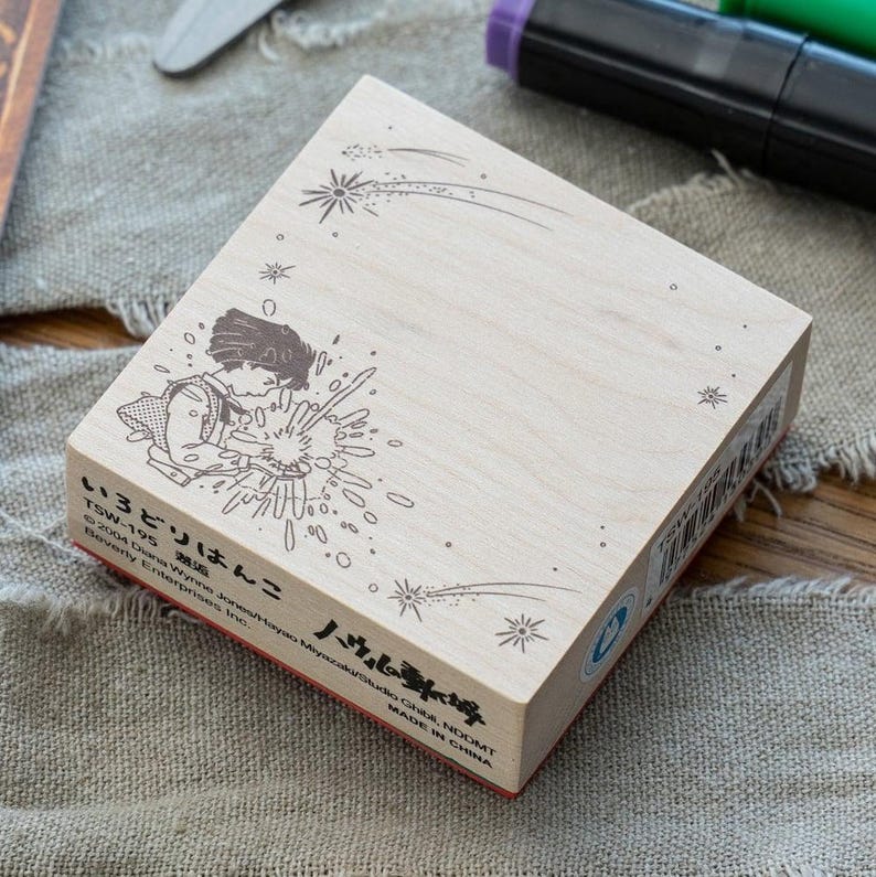 May include: A square wooden rubber stamp featuring a black line art illustration of a character surrounded by starbursts and shooting stars. The side of the stamp includes text reading TSW-195, 2004 Diana Wynne Jones/Hayao Miyazaki/Studio Ghibli, NDDMT.