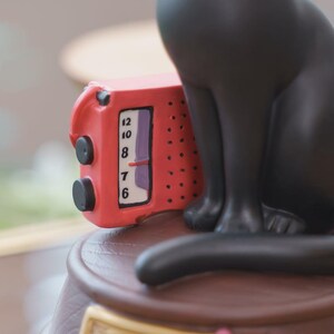 May include: A red miniature radio with a clock face showing the numbers 6, 7, 8, 10, and 12. The radio is part of a black cat figurine. The figurine is sitting on a brown surface.