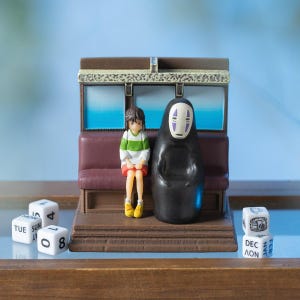 May include: A miniature diorama featuring two figures from the animated film *Spirited Away* on a train car. The figures are Chihiro and No-Face. White dice with calendar dates are in the foreground. The train car has a brown roof and maroon seating.