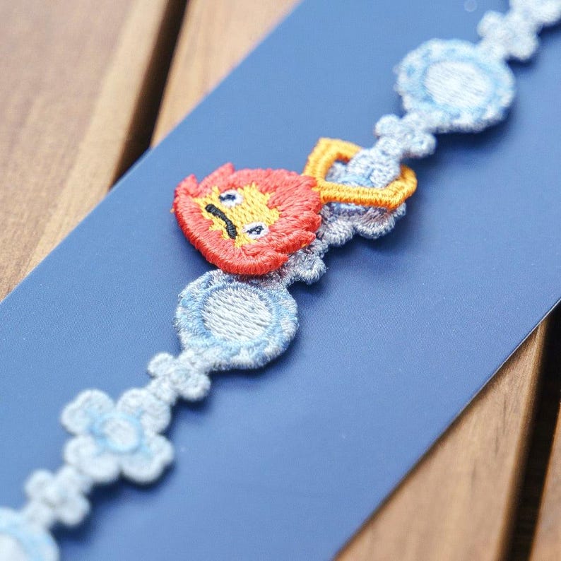 Op de afbeelding: Geborduurde boord met een herhalend patroon van een leeuwenkop en decoratieve vormen in rood, geel en blauw. De boord bevindt zich op een blauwe achtergrond, met een zichtbaar houten oppervlak.